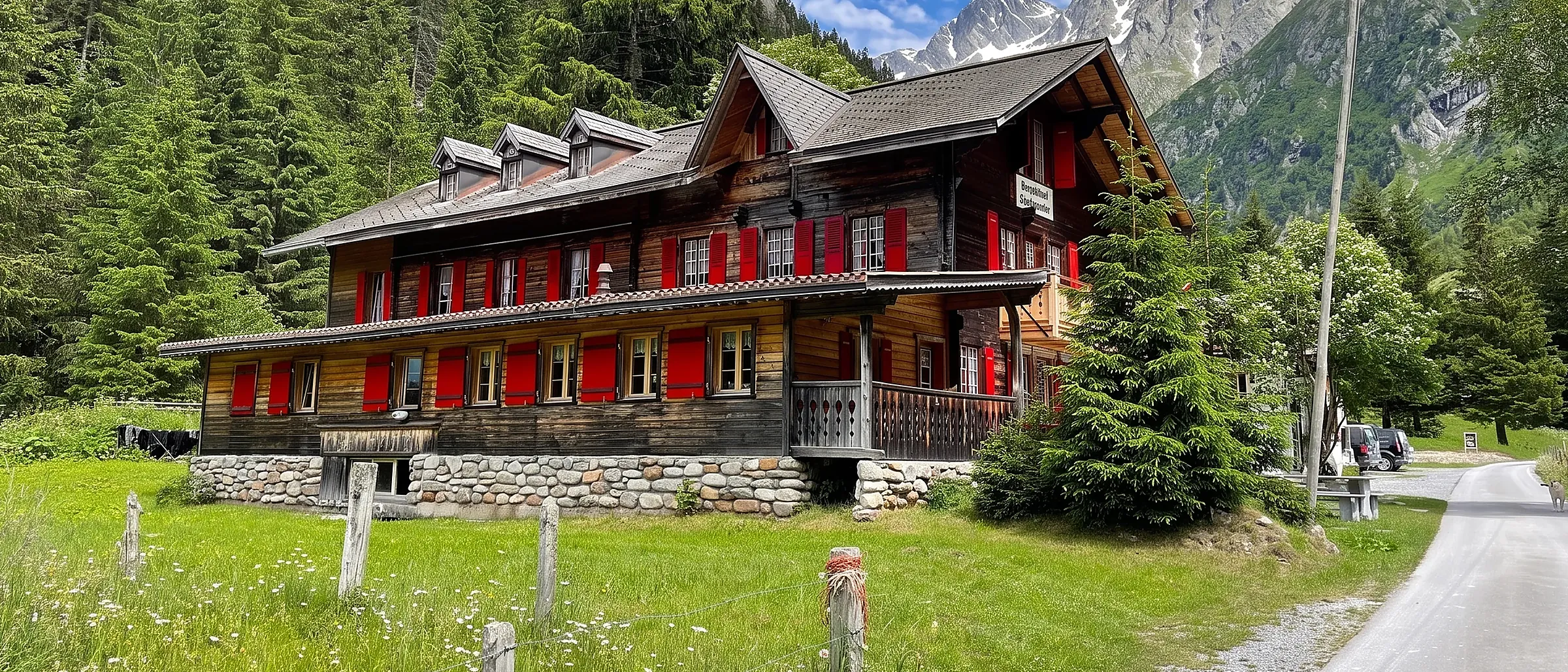 Das Berghotel Gasterntal aus Holz mit roten Läden, davor Wiese und Naturstrasse, dahinter Wald und Fels